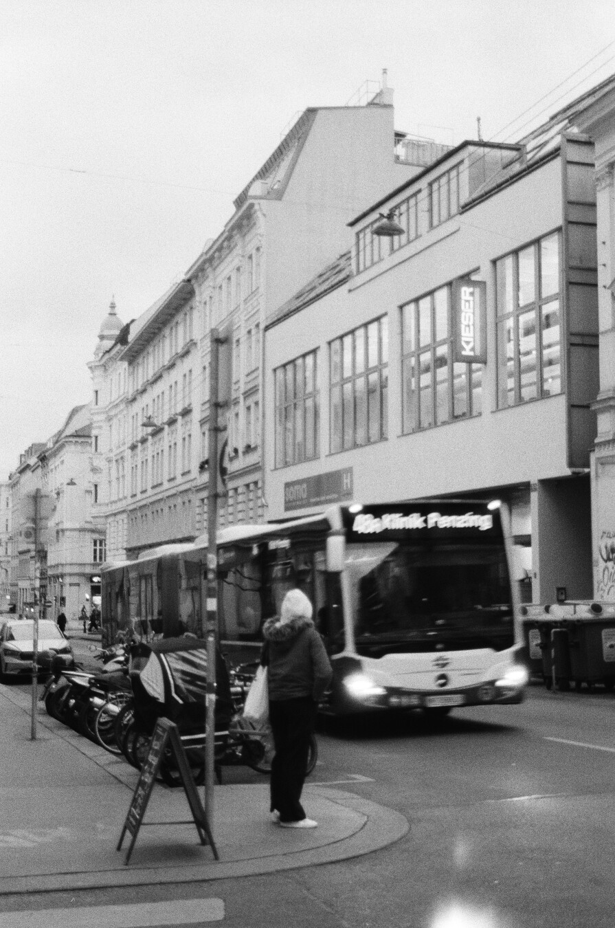 A monochrome photo of a motion blurred bendy bus riding on a street with a pedestrian watching