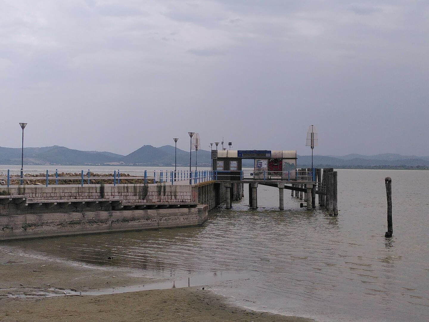 Un molo con ringhiere bianche e azzurre entra nelle acque color sabbia del lago Trasimeno. Lungo la passeggiata vi sono dei lampioni. Sulla parte del molo che guarda la spiaggia c'è una pensilina per l'attesa dei traghetti che è stata scritta e dipinta. Sulla parte destra del molo sono piantati in acqua degli alti pali di legno. Il cielo è grigio. In fondo si vedono le colline.