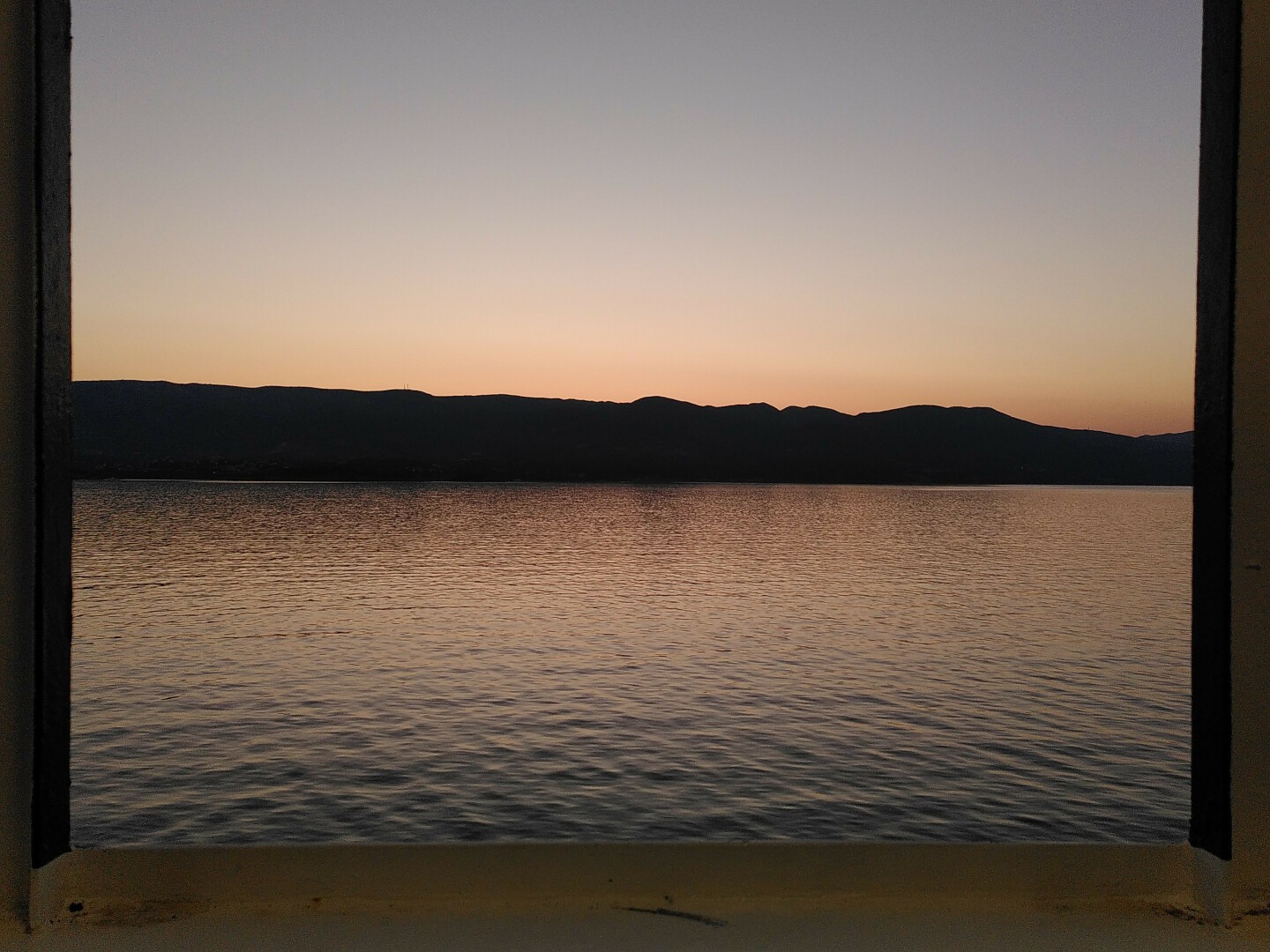 La foto ritrae le prime luci dell'alba viste da una nave. In alto il cielo è azzurro/grigio, e diventa giallo e poi arancione scendendo verso il basso. I colori del mare sono lo specchio di quelli del cielo. Nel mezzo, il profilo scuro di un'isola. La scena è incorniciata negli elementi protettivi del ponte della nave