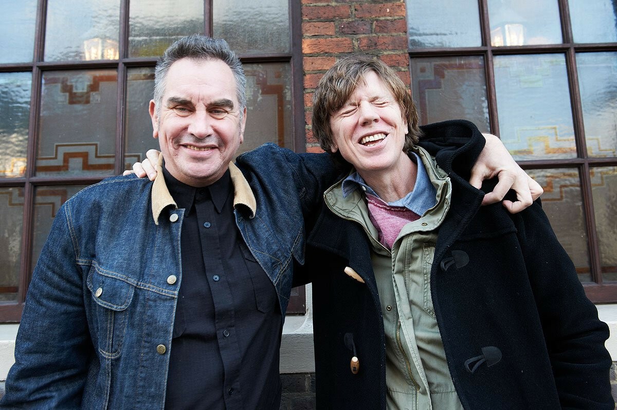 Mark Stewart (a sinistra) e Thurston Moore (a destra) hanno l'uno il braccio sulla spalla dell'altro. Mark sorride, Thurston se la sta proprio ridendo di cuore e ha gli occhi chiusi