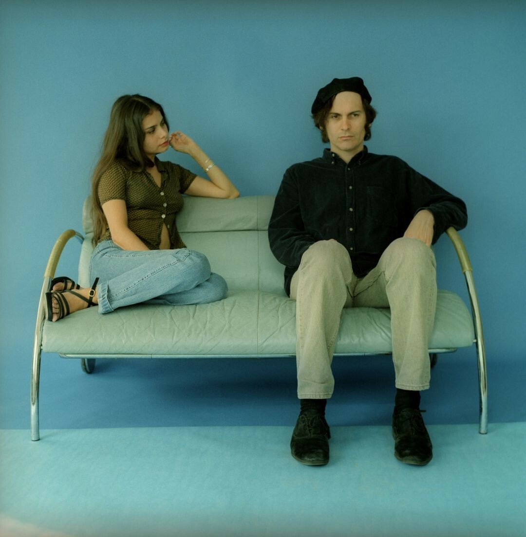 Hope Sandoval and David Roback on a sofa. He's sitting straight, looking somewhere on the bottom left. She's totally on the sofa, turned to the left and looking at Roback. The room is light blue