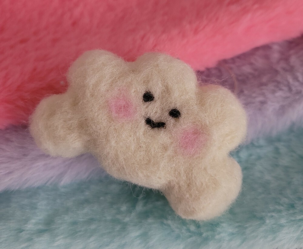 A needle-felted white cloud shape with little cute eyes and smiling mouth, and two pink dots as cheek blushes.