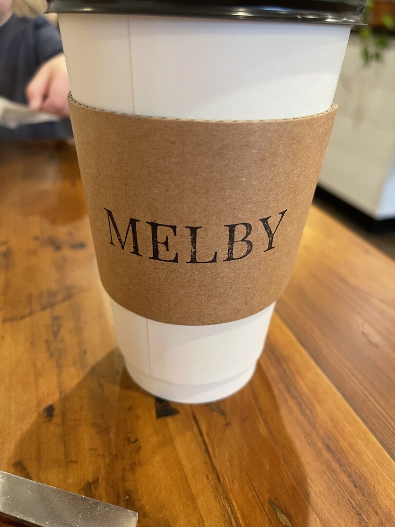 White to go coffee cup with a brown sheath. Black printer letters spelling MELBY, the coffee shop name, on it.