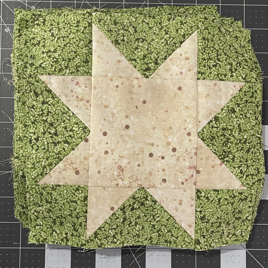 Green and cream star quilting block block on a grey and white cutting mat. Cream star with two points on each side of a center square, pieced together using squares and flying geese.