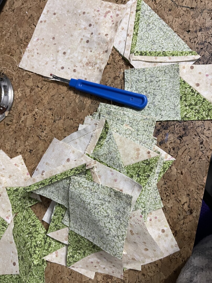 Pieces of a quilt on a cardboard desk. Flying geese blocks made from a cream fabric with brown and mauve dots and a green fabric with a leaf pattern.