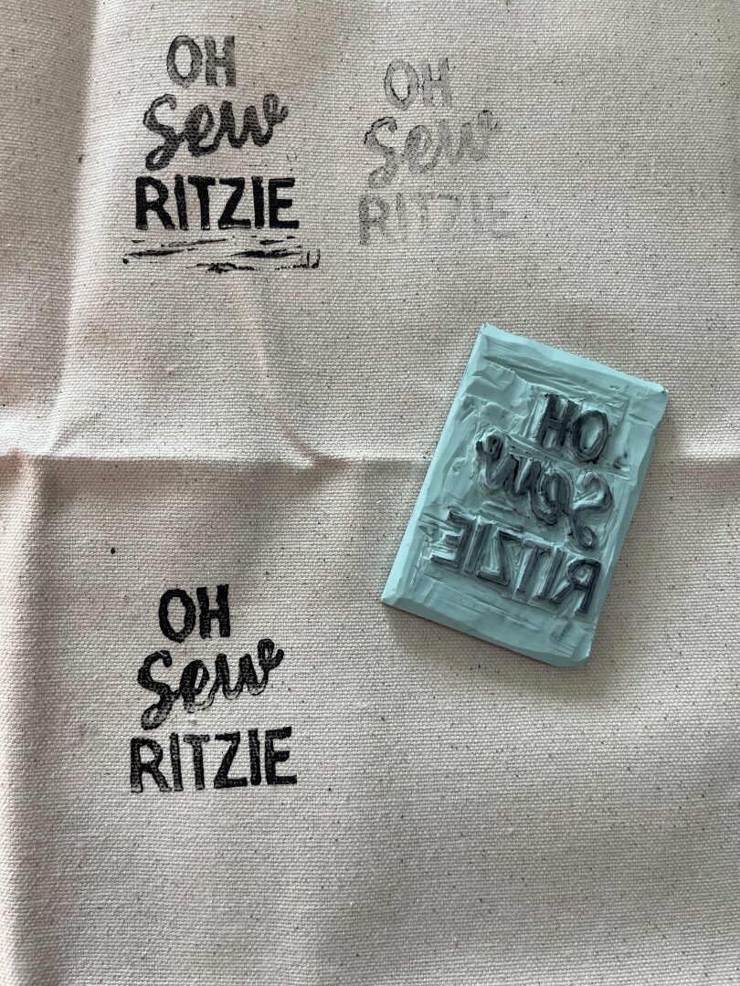 Canvas fabric laid out with three test stamps that say “Oh Sew Ritzie” and the custom stamp I carved.