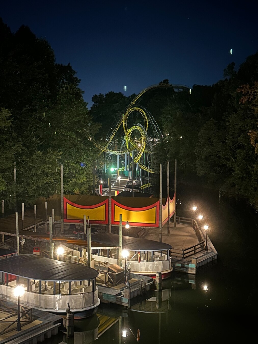 A river scene at night. Two riverboats sit on the river in a well lighted area. A double loop roller coaster is in center of image, also well lighted.