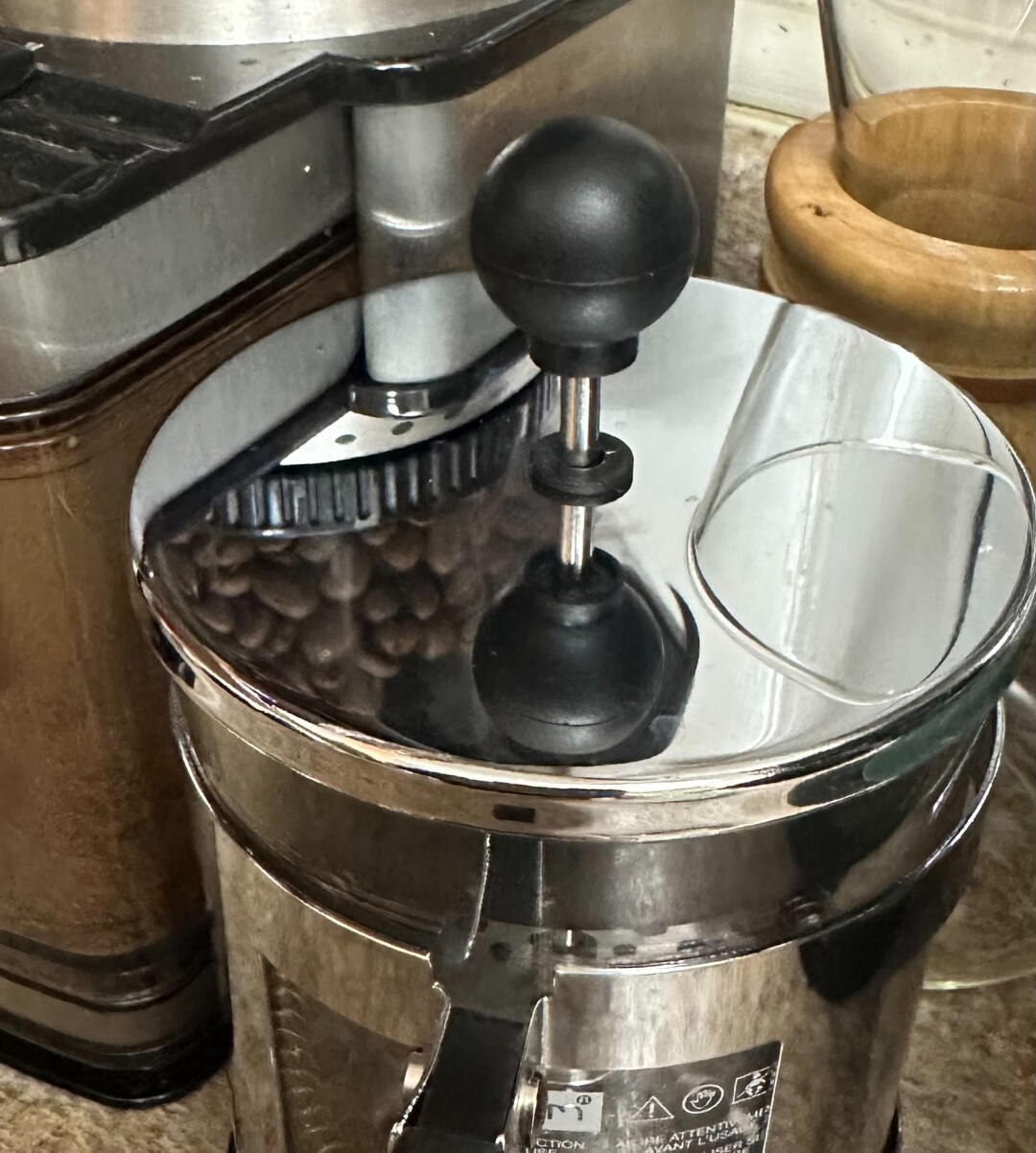 Closeup of the steel lid of a French press reflecting coffee beans from a grinder next to it