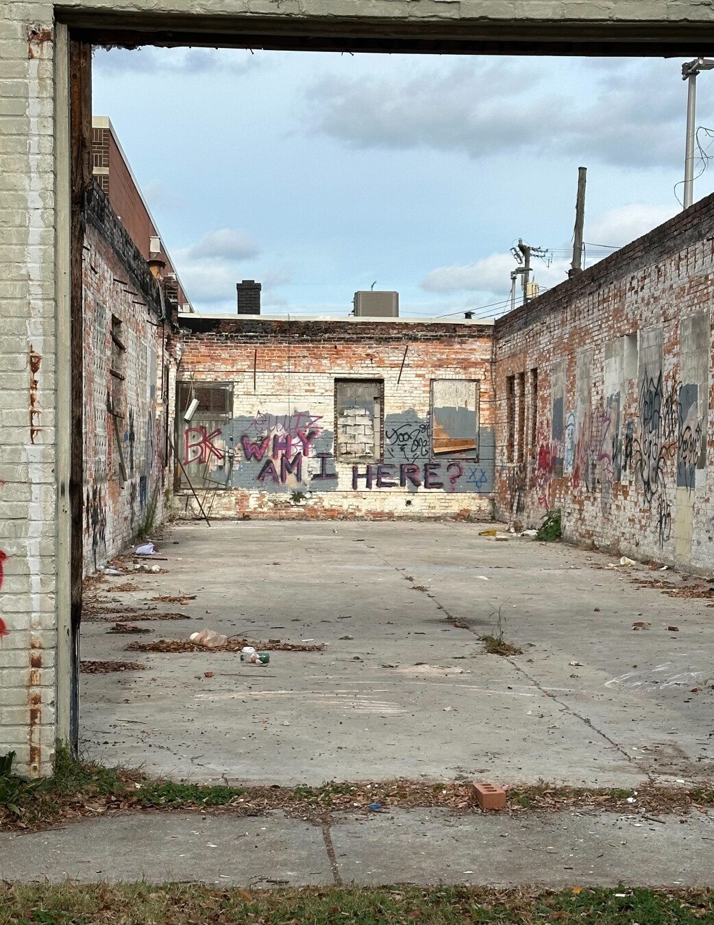 A roofless structure of three walls with faded white paint is covered with various graffiti. On the far wall facing the camera is the text “WHY AM I HERE?” A cloudy sky is visible in the background with a sidewalk and grass in front