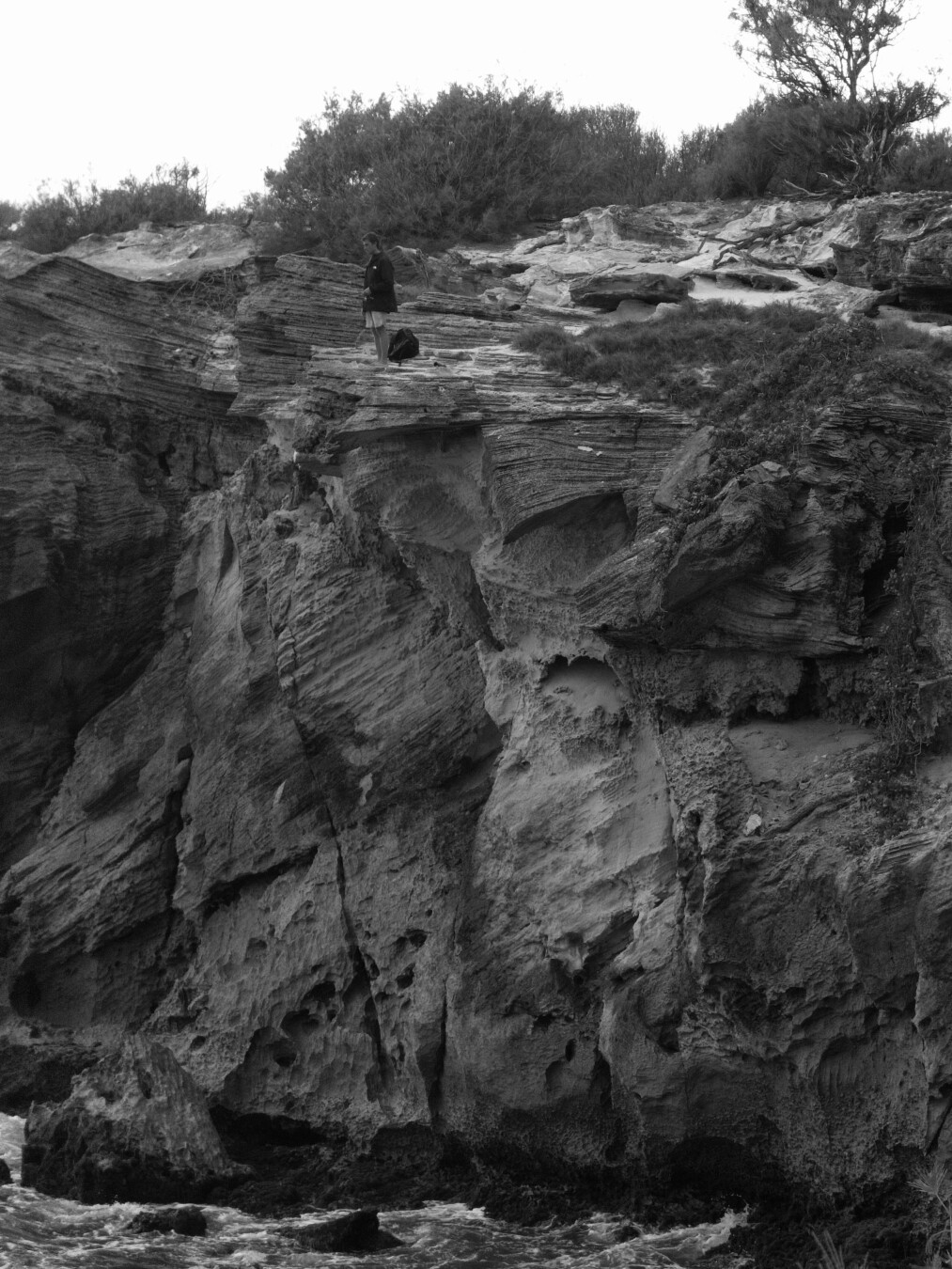 A fisherman waiting for a bite standing at the edge of a large stone cliff over the ocean.