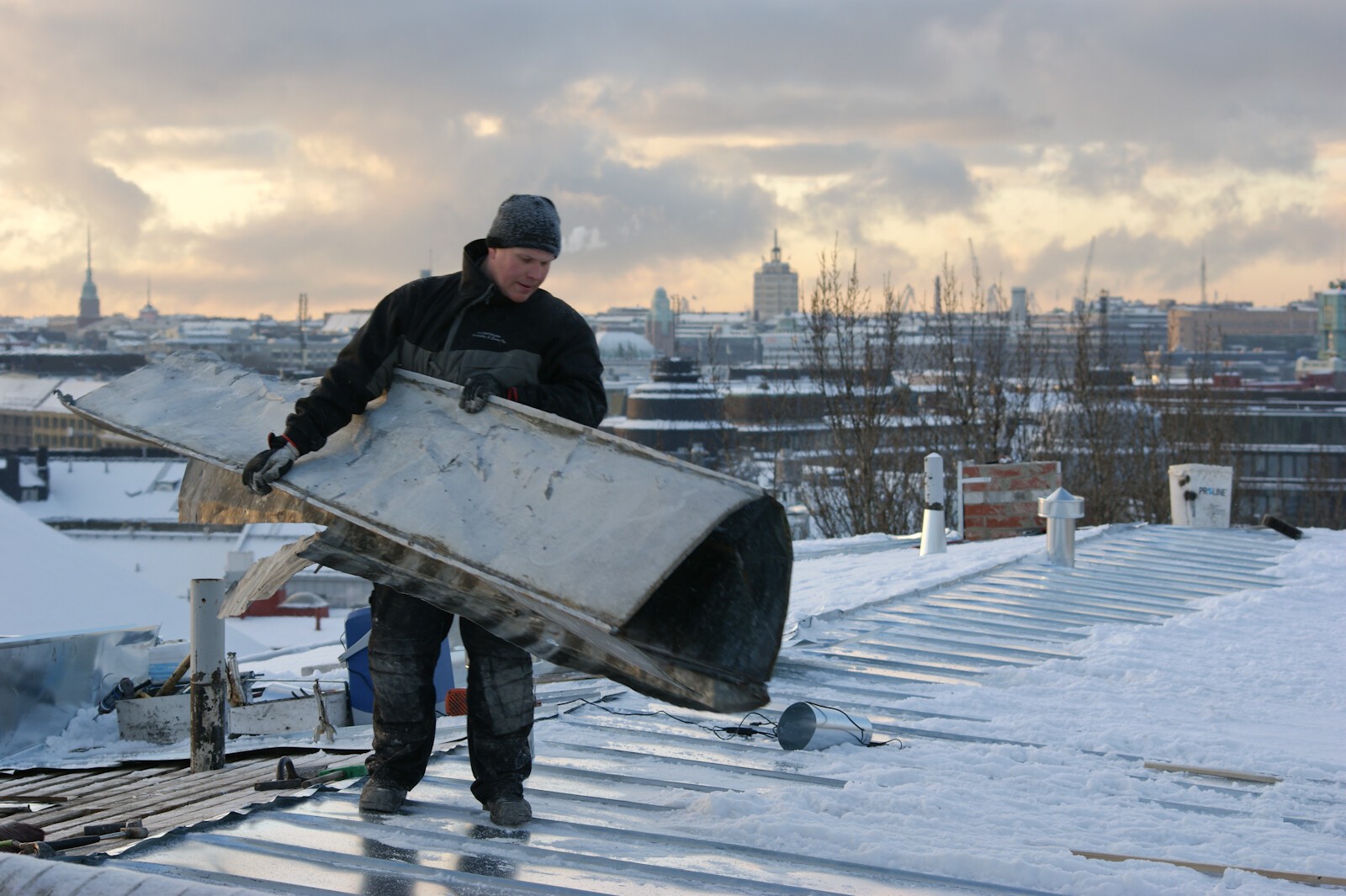 A worker on the snowy roof of a building, on background roofs of a city and clouds in morning light.