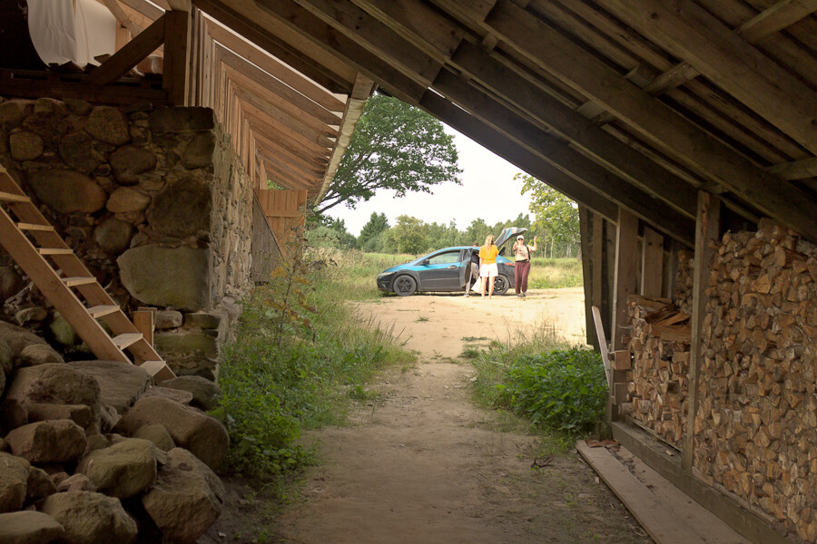 View from under a roof of a building, a new wooden architectural creation on top of stone ruin of an old farmhouse. From an opening between stone wall and roof reaching to ground, one can see to the yard where there is a car and people packing the car to leave.