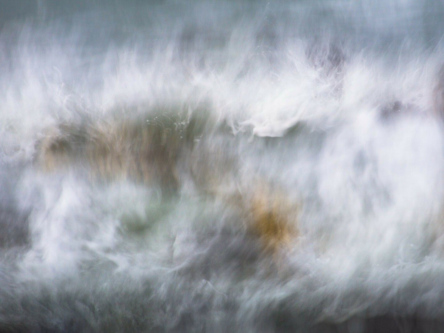 Intentional Camera Movement picture of an ocean wave breaking over rocks. The movement blurs show the dynamism and energy released, with wisps and streak patterns in the foam as it crashes against the rock, mixed in with a few darker brown and black tones of the rock itself. There are blues and greens of the calmer sea in the foreground and behind the breaking wave.