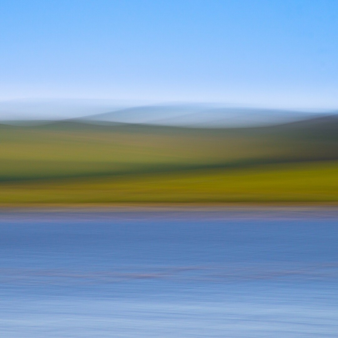 Square colour landscape using intentional movement technique. A pan during exposure of a moorland view on the other side of a lake, with varying green tones of rolling land, darker green lines of gorse and faint after-images recorded. The foreground is deep blue with suggestions of ripples of the lake surface, and a plain blue sky above the moorland.