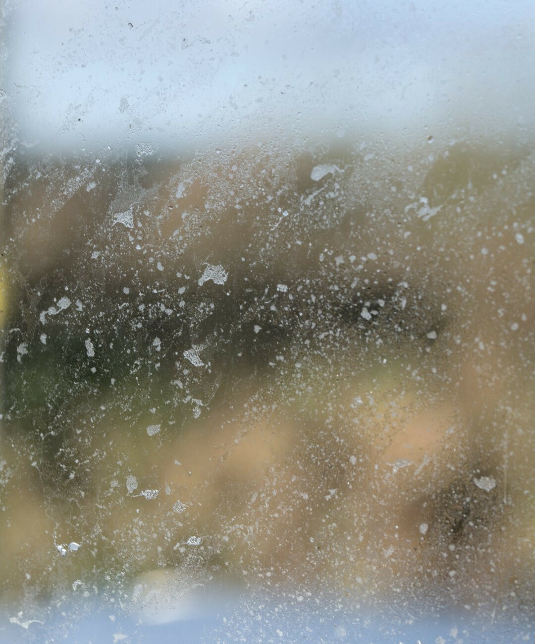 Looking out at a defocused and indistinct view of a landscape, with wintry brown and green tones and a grey colour at the top suggesting sky, through salty rain drops dried onto the surface of a window.