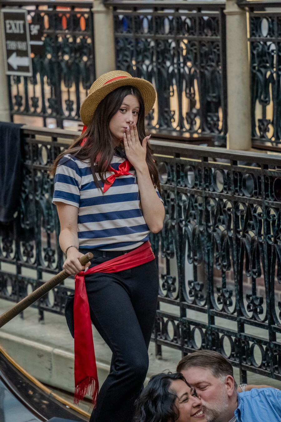 A person dressed as a gondolier stands on what appears to be a boat or platform. They wear a straw hat with a red band, a blue and white striped shirt with a red bow tie, black pants, and a red sash around their waist. The person is holding a long pole, likely a gondola oar, and has one hand raised to their mouth in a comically surprised gesture. Behind them is an ornate metal railing. An "EXIT" sign is visible in the background. In the foreground, at the bottom of the image, two people can be seen partially, a man kissing a woman on the cheek. The setting is in a themed area replicating a Venetian scene. A person dressed as a gondolier stands on what appears to be a boat or platform. They wear a straw hat with a red band, a blue and white striped shirt with a red bow tie, black pants, and a red sash around their waist. The person is holding a long pole, likely a gondola oar, and has one hand raised to their mouth in a comically surprised gesture. Behind them is an ornate metal railing. An "EXIT" sign is visible in the background. In the foreground, at the bottom of the image, two people can be seen partially, a man kissing a woman on the cheek. The setting is in a themed area replicating a Venetian scene.
