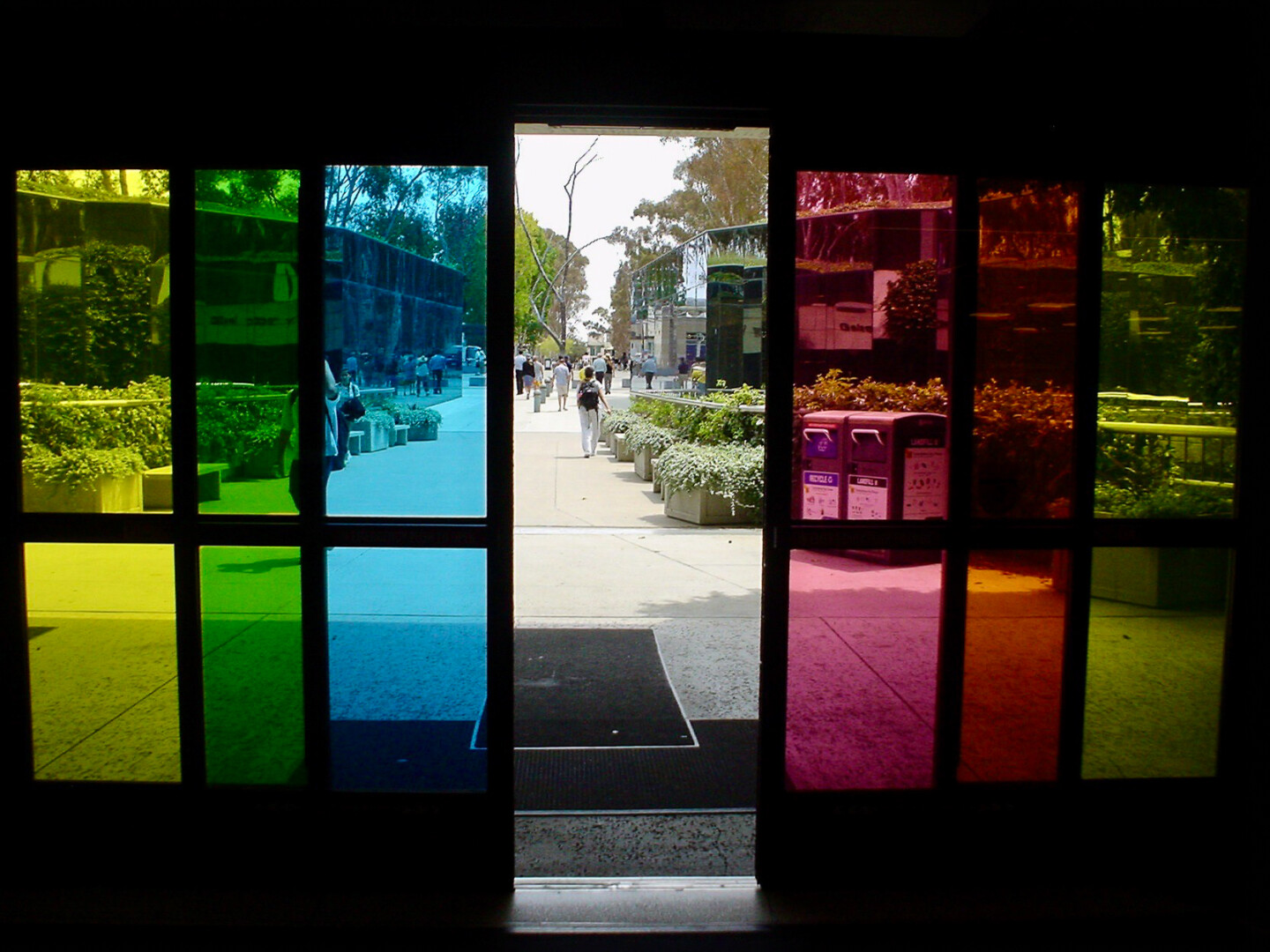 doorway with large glass panels framed in dark material. The glass panels are divided vertically into sections, each featuring vibrant colors: green, blue, yellow, and red. Through these colored panels, an outdoor scene is visible, revealing people walking along a pathway lined with plants and trees. The surrounding area appears sunny and bright, with light reflecting off surfaces.