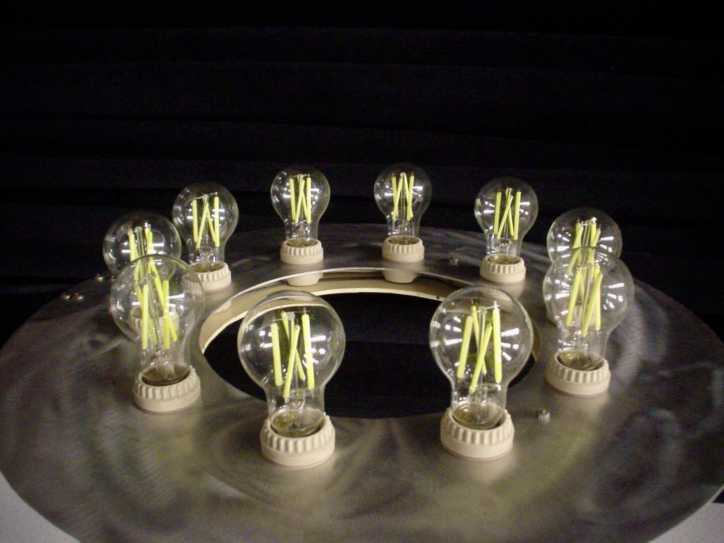A circular metallic base with a brushed, reflective surface. Arranged around the perimeter of the circular base are ten clear light bulbs, each mounted in a beige socket. Inside the bulbs, several yellow matchstick-like objects are visible, standing upright and creating a striking contrast against the transparent glass. The background is a solid black.