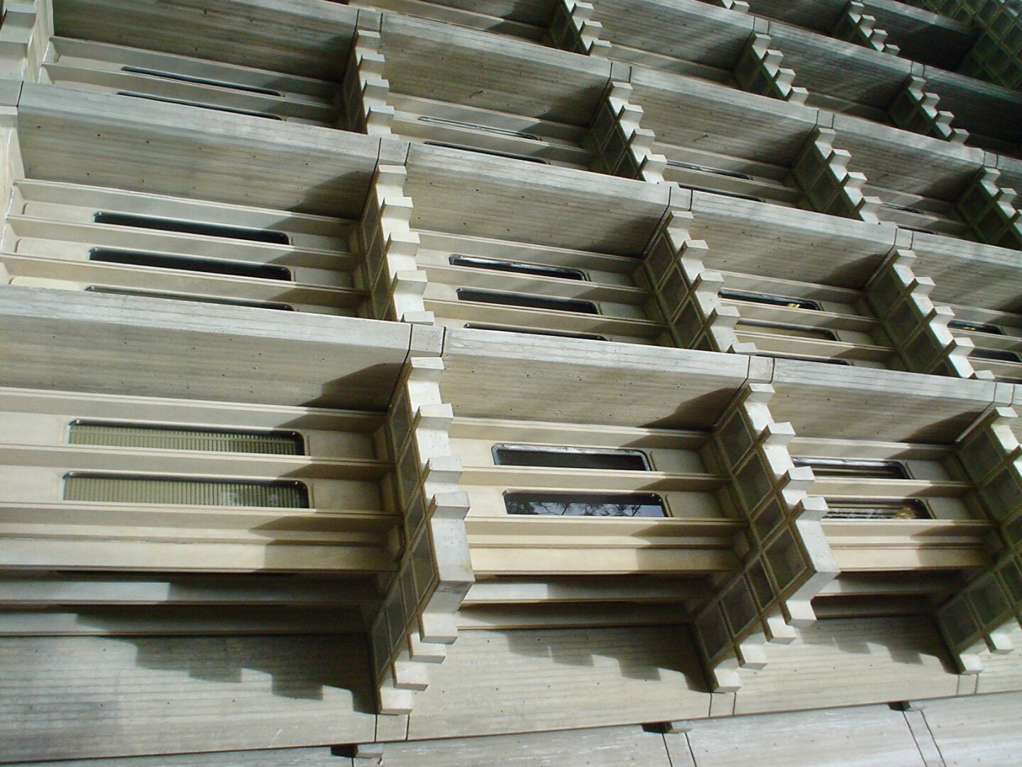 A close-up view of a multi-level architectural structure composed largely of concrete. The design showcases multiple horizontal layers, each with recessed, rectangular windows that provide glimpses of the interior. The individual sections appear to have a modular design, characterized by protruding and indented edges that create a rhythmic pattern across the facade. The texture of the concrete is smooth, with subtle variations in shades of light gray. Some areas reveal reflections, hinting at glass or other material within the windows.