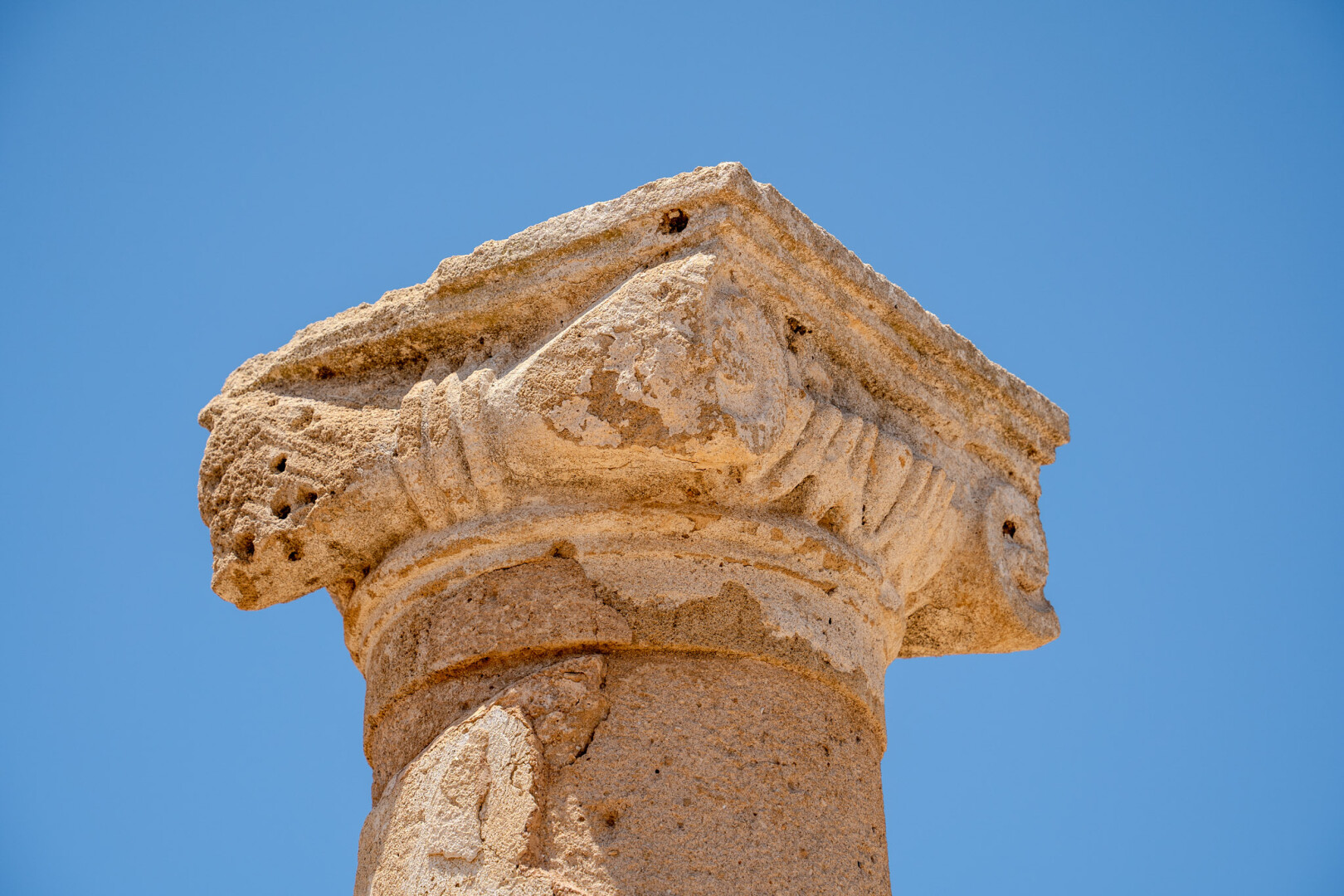 A close-up view of a weathered, tan stone column capital against a clear blue sky. The cylindrical column rises from the bottom of the image into an ornate capital with layered, curved moldings, angular projections, and eroded carved details. The upper section forms a broad, roughly triangular roof-like shape, with chipped edges, pits, cracks, and darker patches from age and weathering.