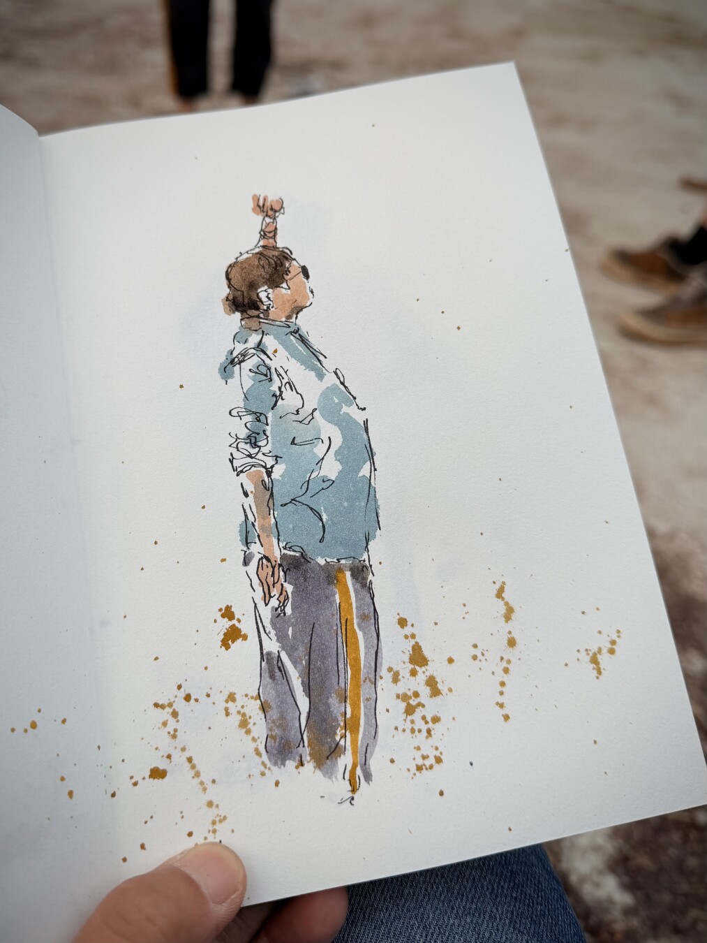 black ink pen drawing filled in with watercolour of someone standing with left arm raised palm facing upwards. Viewed from the right side and right arm towards viewer. Brown hair light blue shirt with short sleeves and grey trousers with orange line. And some orange splashes.