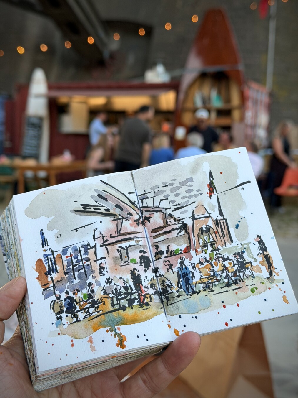holding up my sketchbook in front of scene. Black ink pen with watercolour. A lot of people sitting at tables in front of a food and drink stall. A flag at the right with some red and green, a blue white parasol at the left. And above a part of the metal construction of the bridge. A variety of colours and some watercolour splashes for some texture. It’s comfortable and relax atmosphere