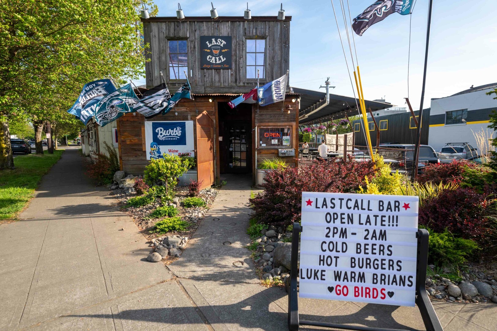 The image depicts the exterior of a lively establishment named "Last Call." The building has a rustic appearance, constructed primarily from wood, with a second-story loft that features two windows. Above the entrance, there is a sign that reads "Last Call" with an illustration of a shot glass and the slogan "Booze Game Fun". The entrance is marked by a wooden door with glass panels, and the place is adorned with various flags and banners, some of which display sports team logos, suggesting a sports bar atmosphere.

In front of the bar, there is a well-maintained garden area with various plants, shrubs, and small rocks. A concrete path with what looks like hoof imprints leads to the bar. There are a few cars parked in the background, indicating that the place might be accessible by road and has a parking area nearby. The setting seems to be in a suburban or semi-urban area with trees visible in the background.

A prominent signboard in front of the establishment provides details about the bar's offerings and operating hours. The sign reads: "Last Call Bar, Open Late!!!, 2 PM - 2 AM, Cold Beers, Hot Burgers, Luke Warm Brians, Go Birds."
