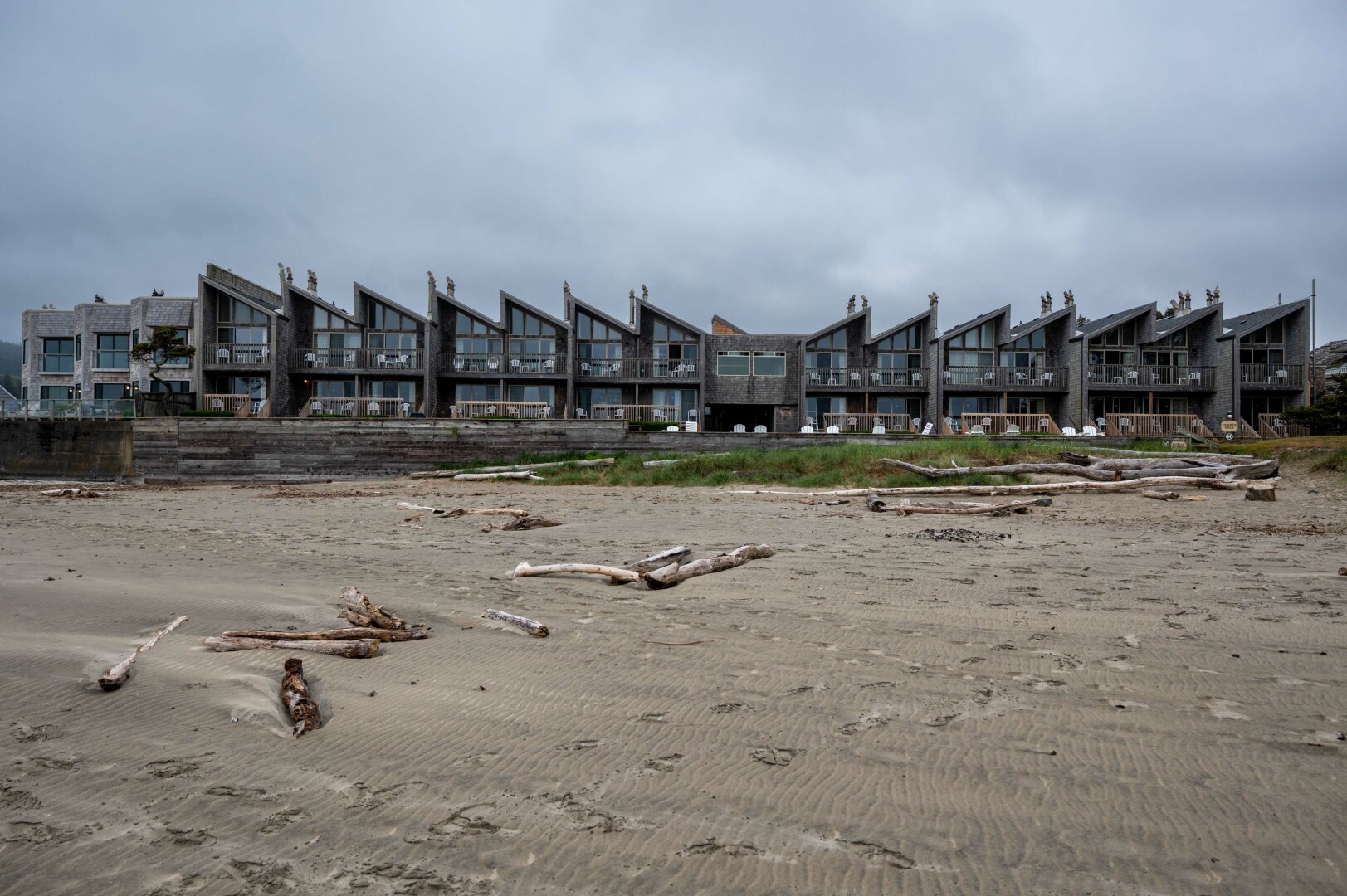 In the foreground—the lower half of the image—there is a sandy beach that appears to be at low tide, as the wet sand is exposed and shows ripples from the receding water. Scattered across the sand are numerous driftwood logs of varying sizes, some of which are bleached by the sun and weathered by the elements. The beach itself seems quiet and unoccupied, with no people visible in the scene.

The sky overhead, which occupies the upper third of the image, is overcast, suggesting a cloudy day with diffused light.

The focal point of the image is a row of a dozen buildings across the middle of the image. Most of the buildings have the same distinct shape with large windows and sliding glass doors that open onto private balconies on two levels, each furnished with a couple of white plastic chairs. The most distincitve design element of these houses are the angled roofs with metal chimneys that create a saw-like overall shape that runs across the width of the image.