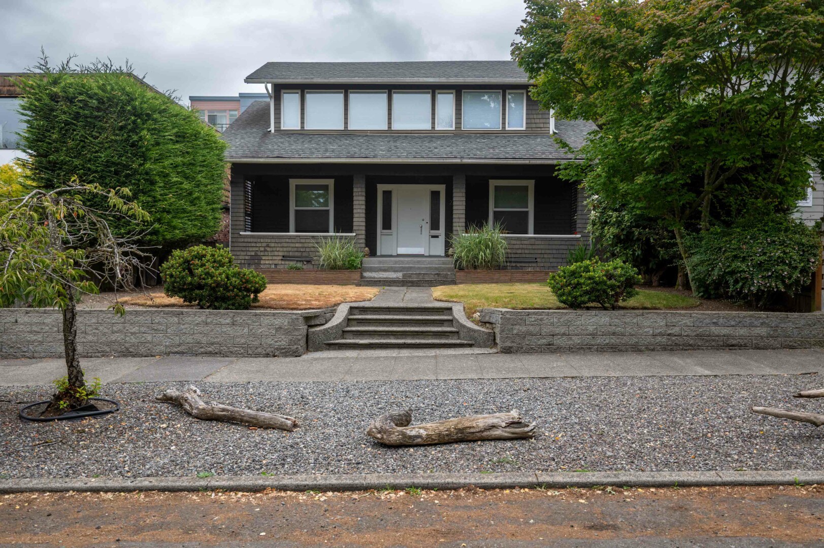 The image depicts a two-story house with a sleek and contemporary design. The house is constructed primarily of dark materials, including dark wood shingles, giving it a sturdy and elegant appearance. The front of the house features a central entrance with a white door, flanked by two windows on either side. Above the entrance, on the second level, there is is a series of seven tightly spaced windows that spans the width of the house, allowing ample natural light to enter the interior.
In front of the house, there is a small set of stone steps leading up to the entrance, which is elevated slightly above ground level. The steps are wide and appear to be made of the same stone material as the surrounding walls and pathways. The area immediately surrounding the house is well-maintained, with a combination of greenery and hardscaping. There are bushes and small trees planted around the perimeter, adding a touch of nature to the otherwise modern aesthetic.
The area in front of the house between the sidewalk and the street is made of gravel, providing a rustic contrast to the polished look of the house. There are a few large logs lying on the gravel, possibly for decorative purposes or to add a natural element to the landscape. The sky above is overcast, indicating a cloudy day.
