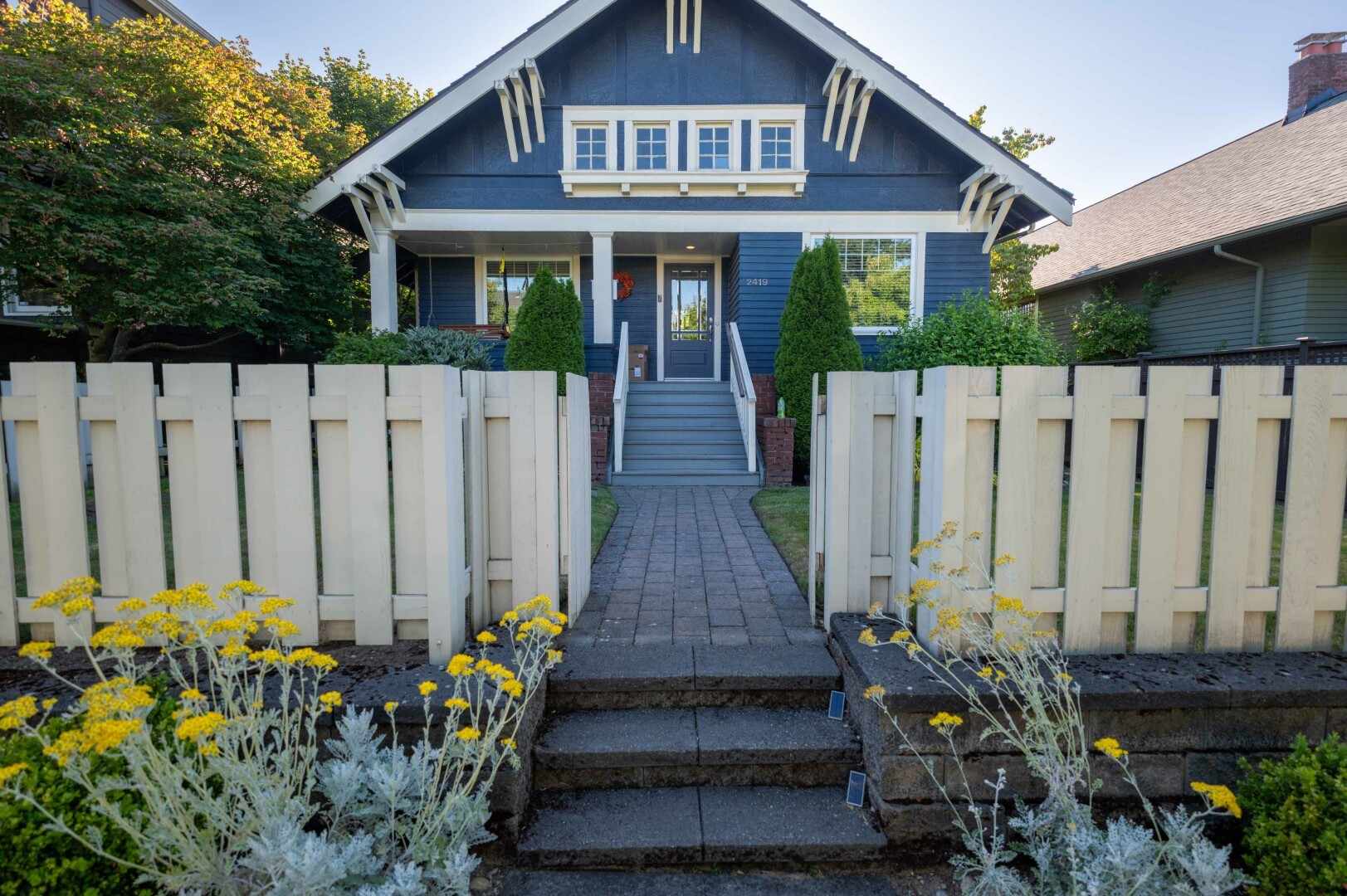 This image depicts a charming house with a well-maintained front yard. The house is painted in a deep blue color with white trim, giving it a classic and inviting appearance. The front entrance features a set of stairs leading up to a small porch, flanked by two neatly trimmed coniferous shrubs. The house number, 2419, is visible on the right side of the door. The overall architecture suggests a traditional style, possibly from the early 20th century.
The front yard is enclosed by a white picket fence, adding to the quaint and picturesque feel of the scene. The fence is composed of evenly spaced vertical slats, creating a sense of order and symmetry. In the foreground, there are flower beds filled with yellow flowers and some greenery, adding a touch of nature and color to the scene. The pathway leading to the house is made of stone or brick, providing a sturdy and attractive walkway.
The lighting in the image suggests it was taken during the late afternoon or early evening. The sunlight casts a warm, golden glow on the house and the surrounding area, enhancing the colors and creating a cozy atmosphere. The shadows are long and soft, indicating the sun is low in the sky. The composition of the photo is well-balanced, with the house as the central focus, the fence and flowers framing the scene, and the pathway drawing the viewer's eye towards the entrance.