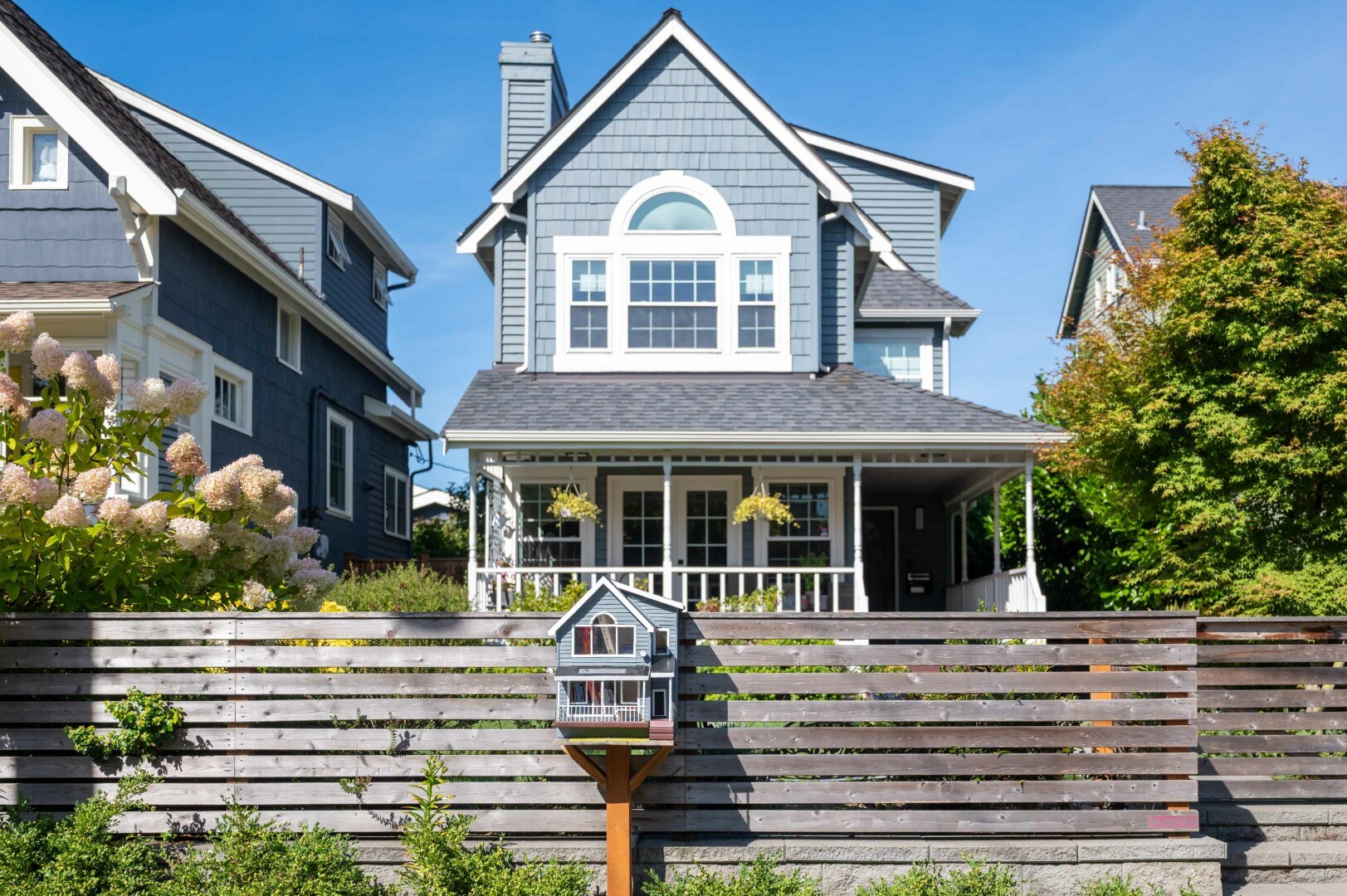 This image depicts a charming residential scene on a bright, sunny day. The focal point is a large, two-story house with a steeply pitched roof and a prominent gable window at the top. The house is painted in a medium shade of gray with white trim. The front porch features white railings and columns, adding to the house's traditional aesthetic. The house is surrounded by lush greenery, including a well-maintained lawn and various plants, which contribute to the serene and picturesque setting.
In the foreground, there is a wooden fence that runs horizontally across the image. On top of this fence, there is a Little Free Library that is a smaller replica of the main house. The lighting in the image is bright and natural, suggesting that the photo was taken during the daytime under clear skies.