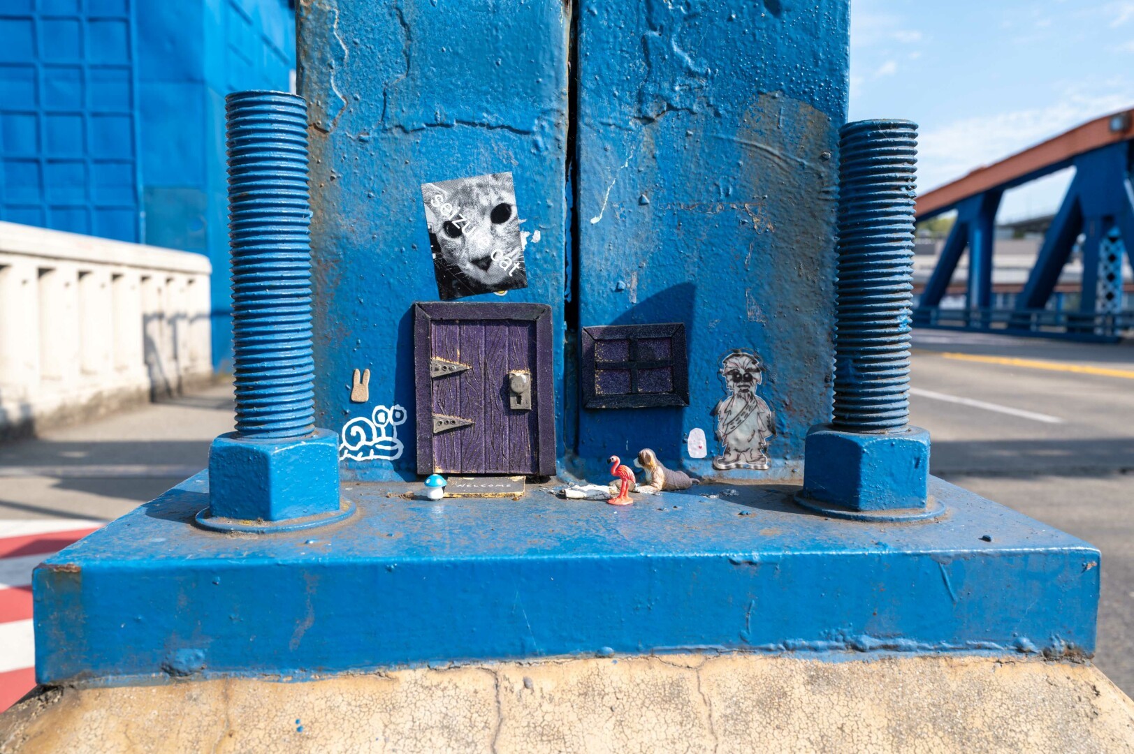 This image captures a whimsical and artistic scene set against the backdrop of a bridge. The focal point is a blue-painted metal structure, likely part of the bridge's support system, adorned with various small, creative elements. The structure features two large bolts on either side, adding to its industrial feel. The blue paint is weathered and peeling in places, revealing patches of rust and the original metal underneath, which adds a layer of texture and history to the scene.
The artistic elements on the structure include a small wooden door with two golden arrow knobs, giving the impression of a tiny house. Above the door is a sticker of a cat's face, adding a playful touch. To the right of the door, there is a small figurine of a person, and below it, two small animal figures, one red and one gray, appear to be interacting. These elements create a sense of a miniature world or a hidden scene within the larger industrial setting.
The lighting in the image is bright and natural, suggesting it was taken during the day. The sunlight casts soft shadows, particularly noticeable on the right side of the structure, indicating that the sun is likely positioned to the left. The background includes a blue bridge and a clear sky, providing a contrast to the detailed foreground. The composition of the photo is well-balanced, with the blue structure and its artistic elements centrally placed, drawing the viewer's attention immediately to the creative details. The background elements, such as the bridge and the sky, frame the scene and provide context.