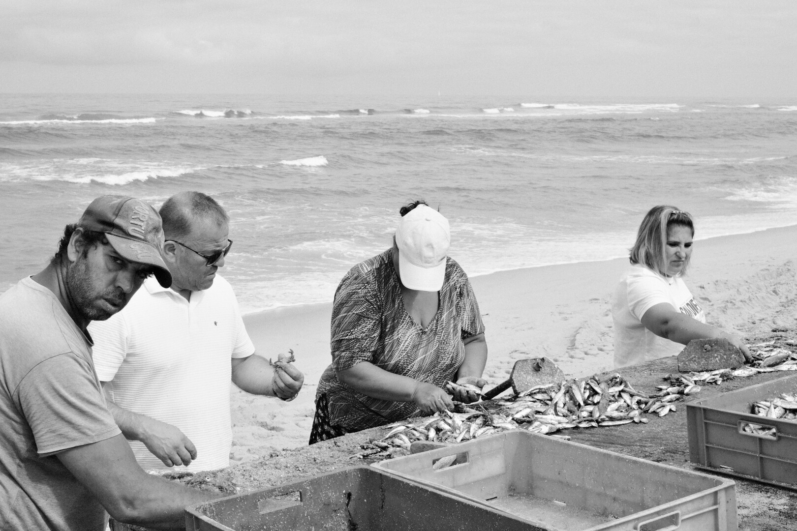 BW picture of a couple people sorting fish on a wooden table the fishermen on the left watches me taking the picture. In the background the sea.