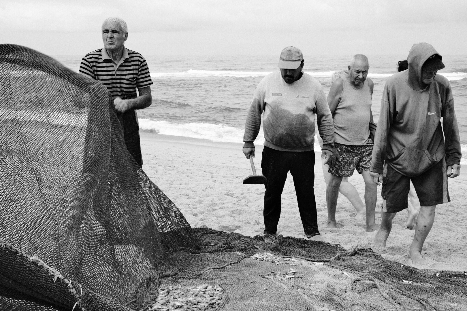 BW picture of a couple fishermen cleaning up a net. On the left one of them is holding the net up, and next to him stands another fishermen with a wooden tool, looking at the net.