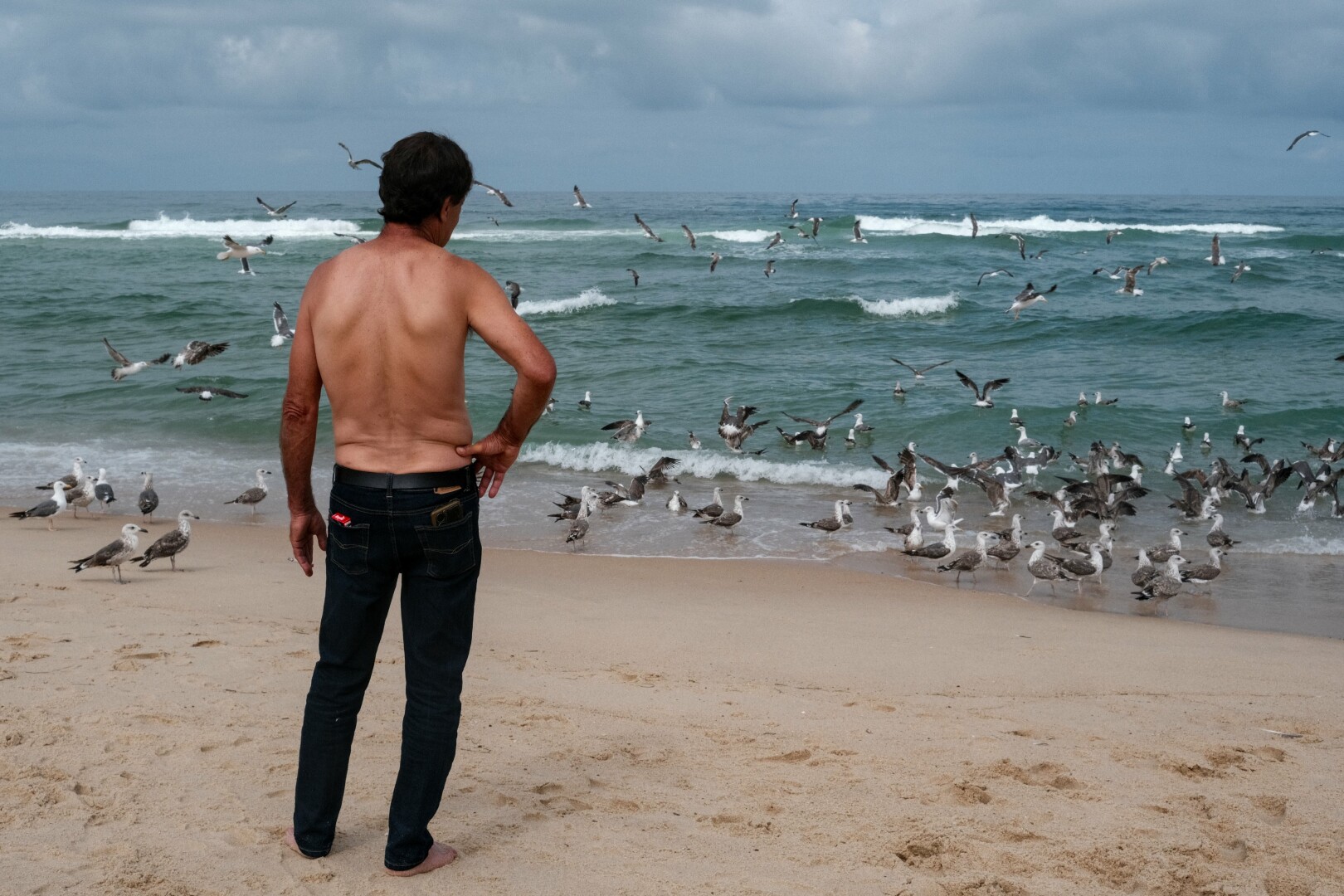Picture of a man, seen from behind, who is having a smoke while looking at the sea. He is shirtless wearing and dark blue jeans and has a pack of cigarettes in his backpocket. On his upper body the tan line from wearing a tshirt is visible. On the right side a bunch of doves can be seen.