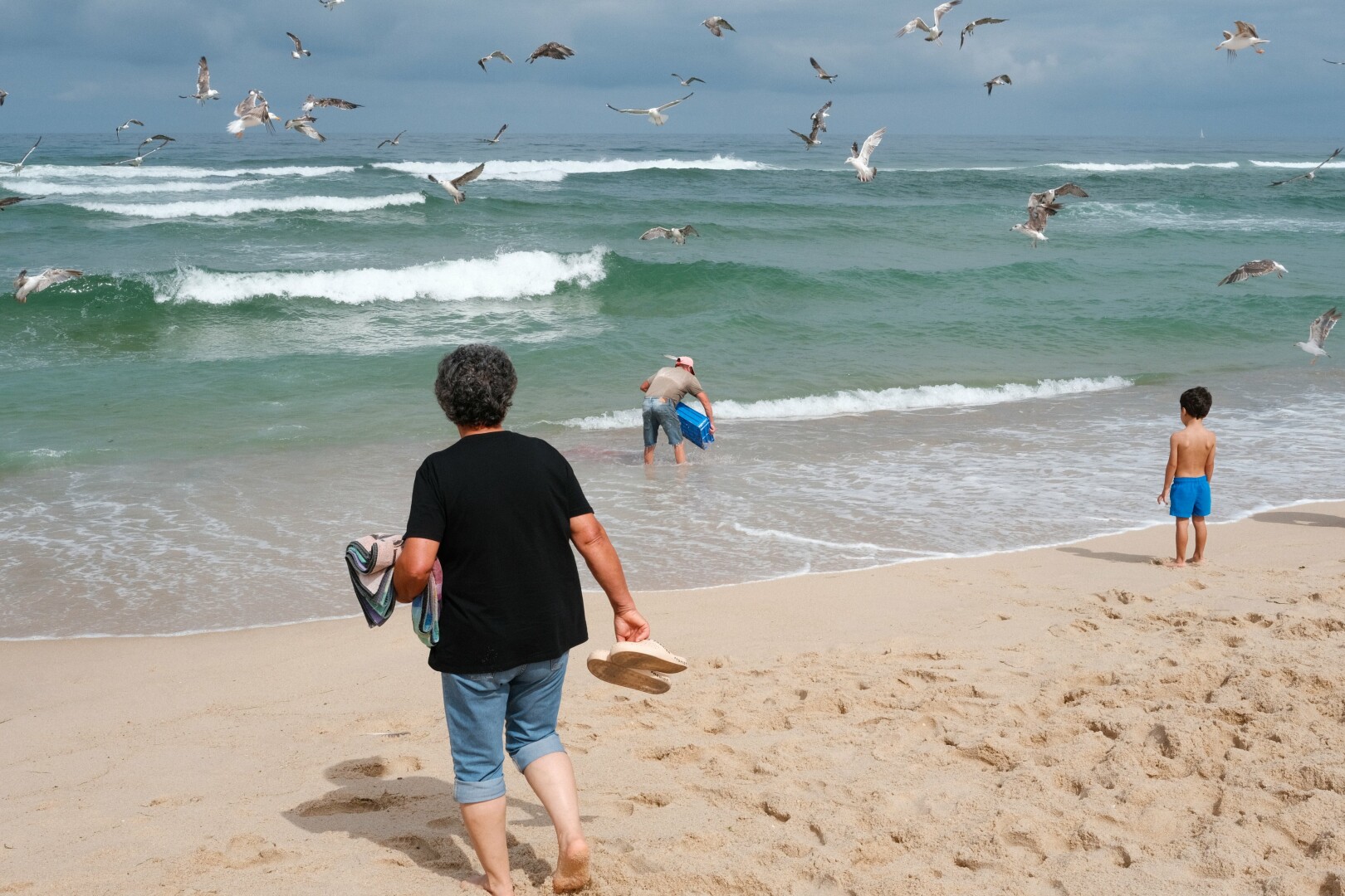 BW picture of a single fishermen cleaning a plastic box using the seawater. He is surrounded by doves trying to find some leftovers in the water. On the beach, to the right, a little boy is watching the scene, and in the foreground the mother watches the boy.