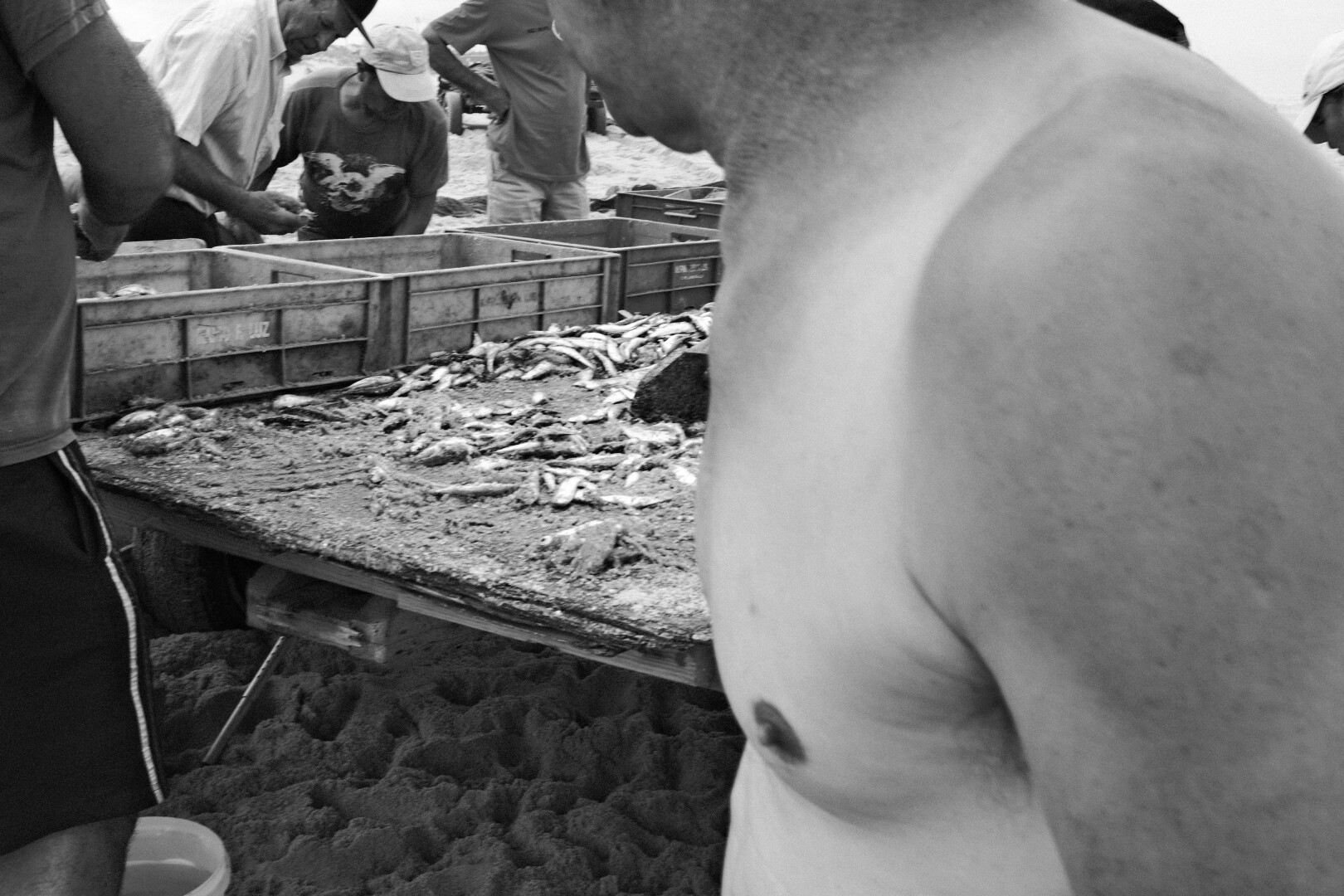 BW picture of the wooden table where the fish is sorted. Upfront from the right, a fishermen without shirt walks into the picture.