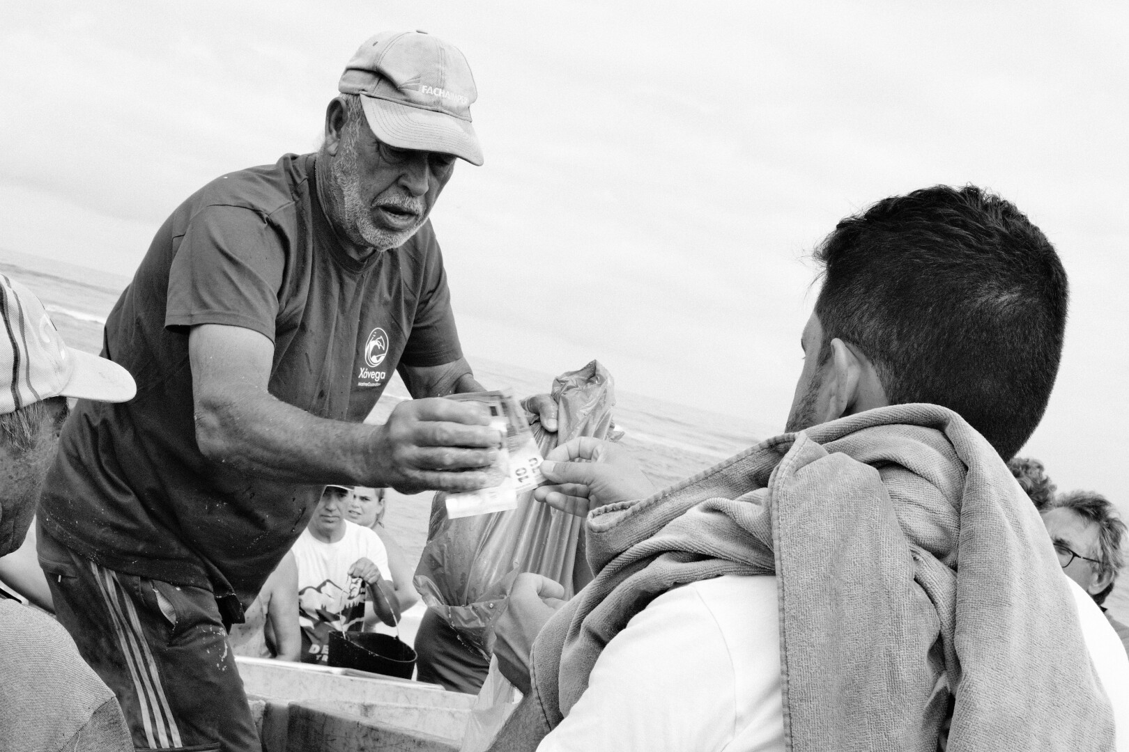 BW picture of the interaction between a fishermen and a client with a beach towel on his shoulder: the client is receiving a plastic bag with fish and handling over some money.