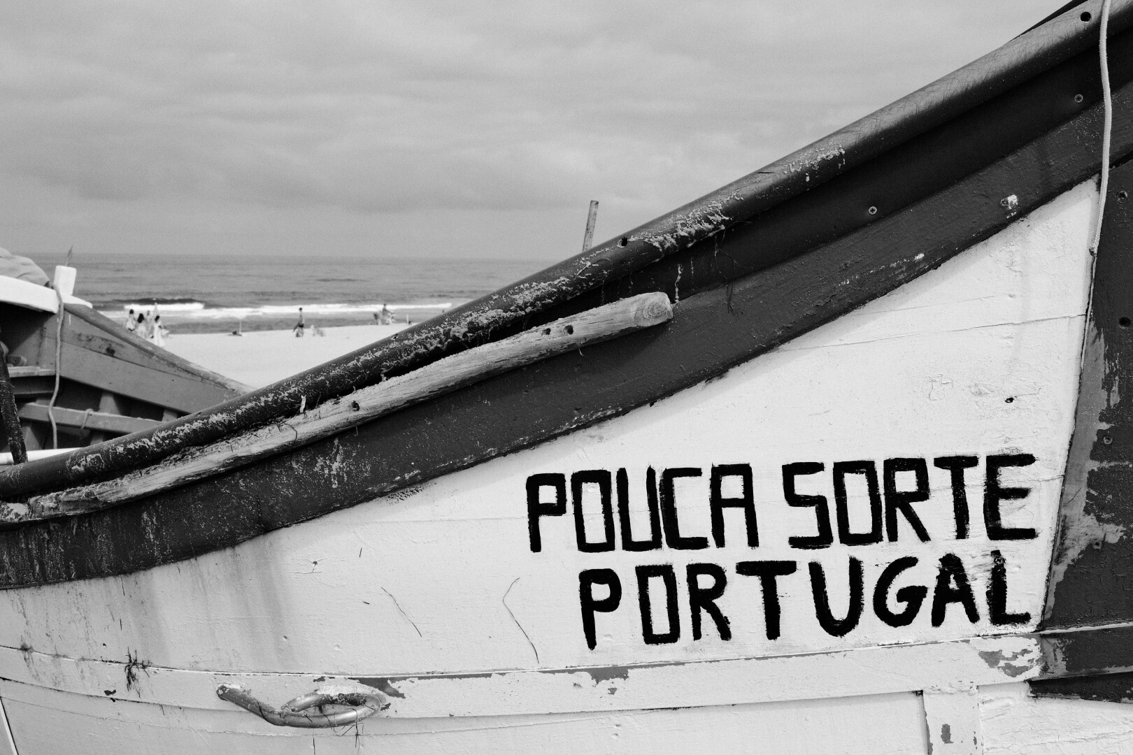 BW picture of a small, wooden Fisher boat.  Upfront the name and country: Pouca Sorte (Bad Luck) Portugal. In the background the ocean is visible.