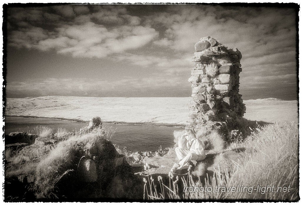 A toned black and white infrared photo of a ruined stone building above a sea inlet, partly overgrown. A young girl is sitting down, shading her eyes as she looks out to sea.