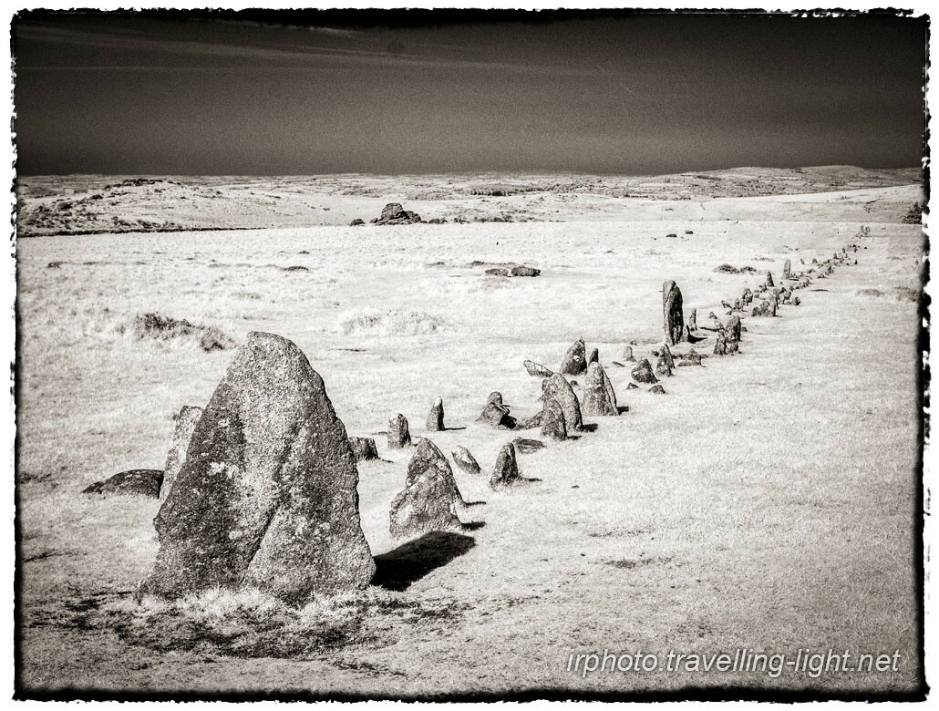 A toned black and white infrared photo of moorland with a standing stone and a double row of smaller stones stretching away.