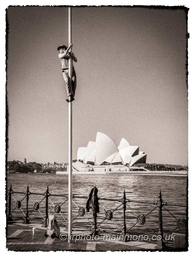 A toned black and white infrared photo of a man who has climbed a flagpole on a pier and is looking out to the left of the frame. There is an iron railing at the edge of the pier and the Sydney Opera House is in the background.