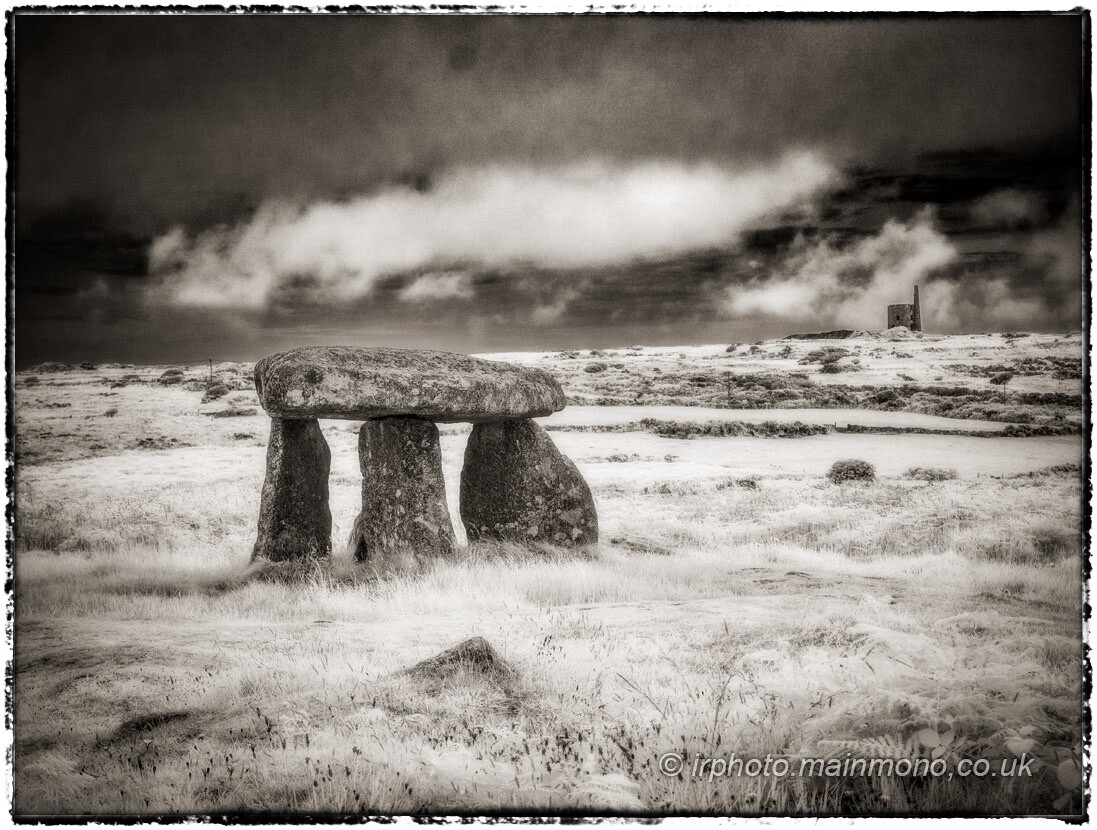 A toned black and white infrared photo of a prehistoric burial chamber or dolmen, consisting of three standing stones supporting a flat capstone, standing in open moorland. There is a ruined mine engine-house on the horizonj