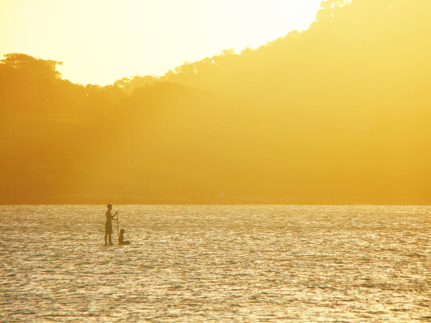 picture at sunset of a man standing and a little kid sitting on a standup board at the distance.