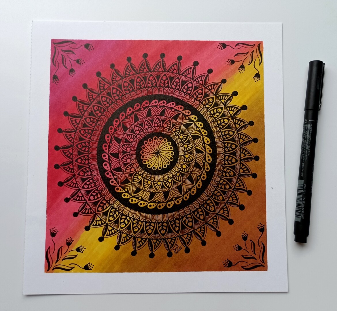 A photo of a black mandala drawing on a paper with red and brown gradient background and flowers in the corners.