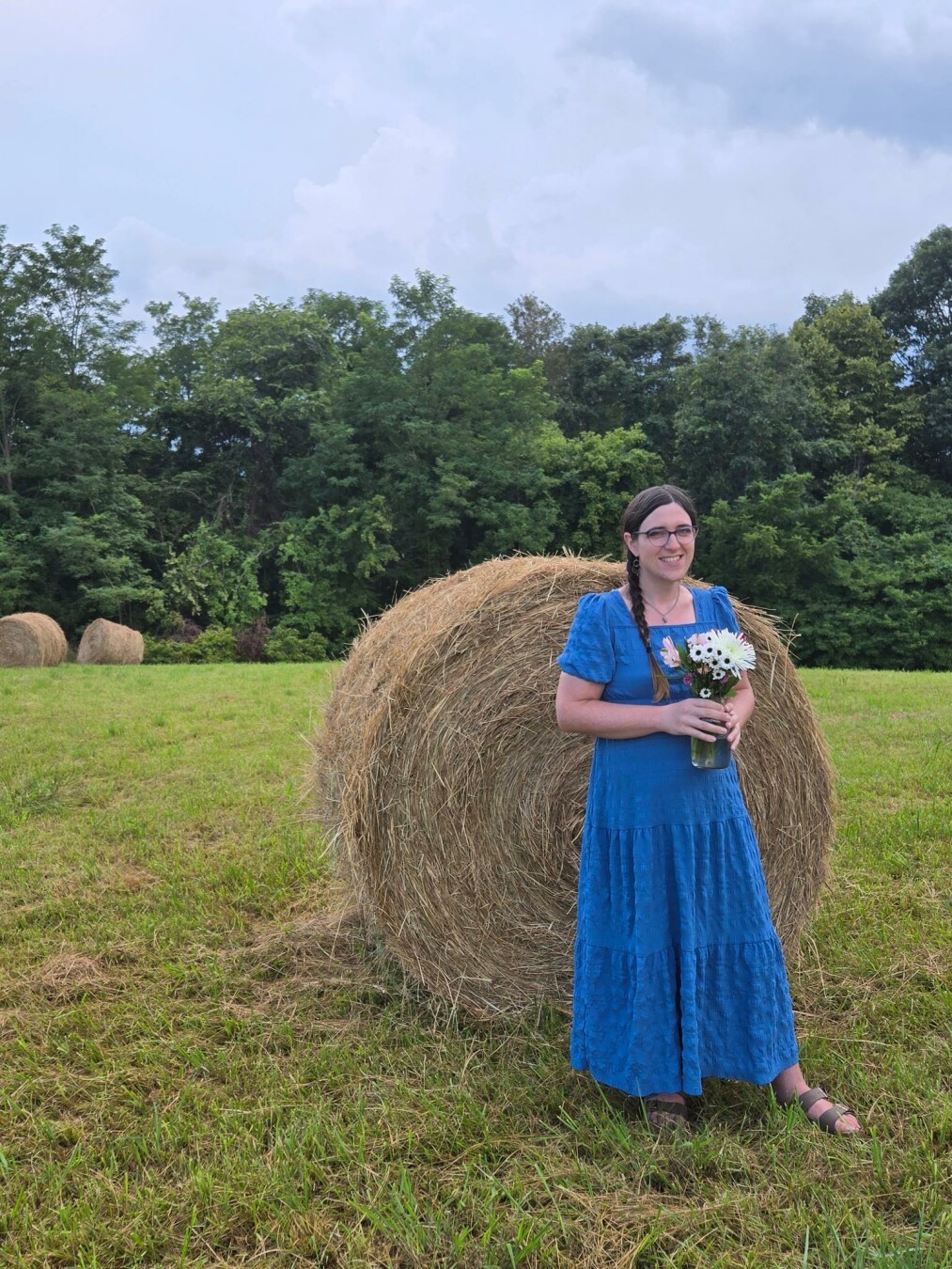 Wide shot of white trans woman in a blue dress with a braid holding flowers in front of a hay bale.