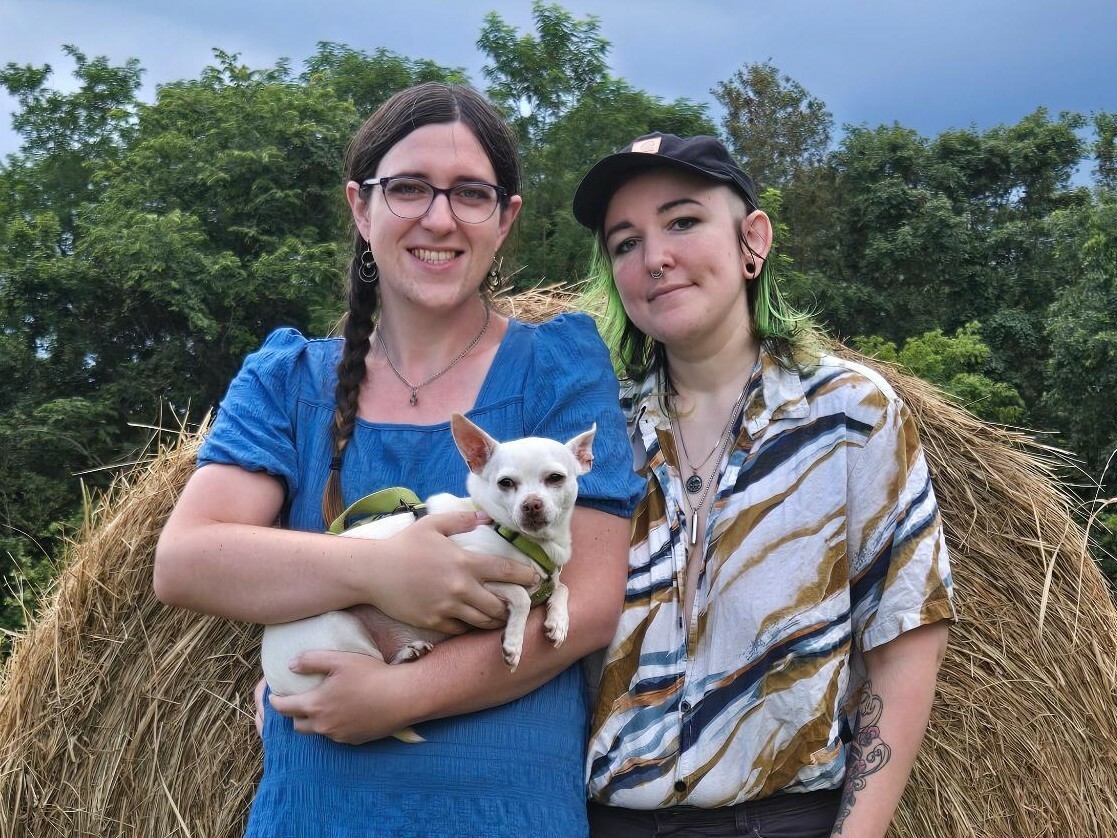 White trans woman with a braid in a blue dress holding a small white Chihuahua and a white person with green hair, ball cap, and light shirt stand in front of a hay bale.