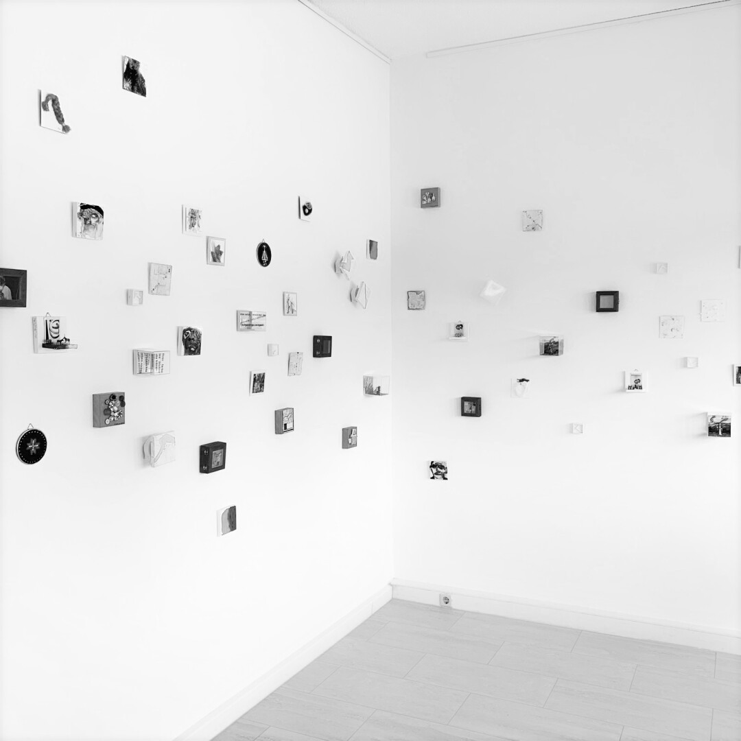 Quadratisches schwarzweißes Bild einer Raumecke. Es hängen viele kleine Werke an den weißen Wänden.