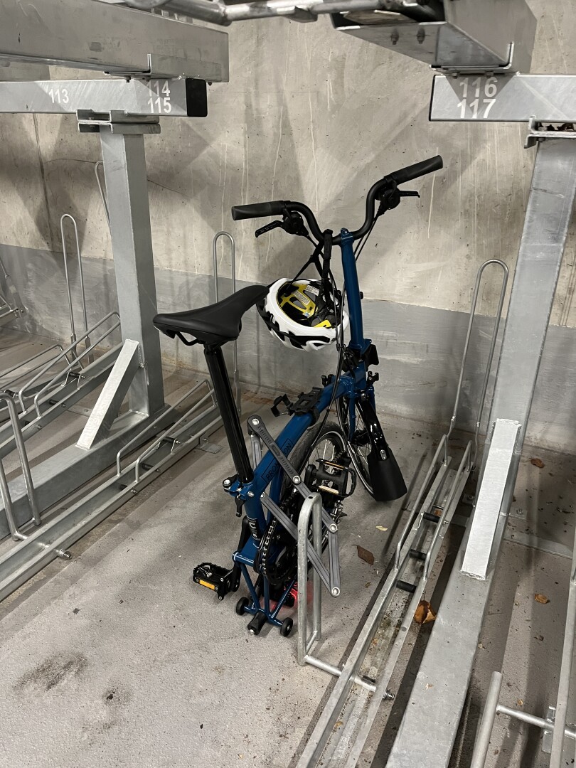 Ein halb gefaltetes Faltrad mit Helm dranhängend in einer Lücke zwischen zwei doppelstöckigen Fahrradständern.
Vielleicht denkt es sich, gut, dass es hier someone Lücke für mich gibt, ich bin noch zu klein für den Pro Ständer…