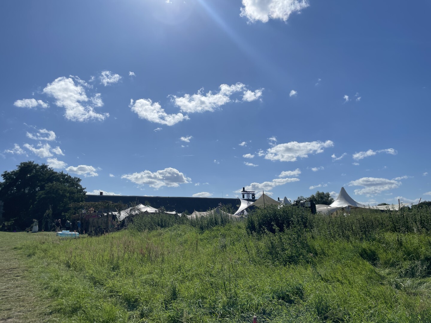 Foto einer grünen Wiese, im Hintergrund sieht man Spitzen von weißen Zelten, ganz Link ein grüner Baum mit runder Krone. Die Sonne ist oben grad nicht mehr im Bild aber man sieht einige Strahlen, die auch den blauen Himmel und Federwolken anstrahlen.
